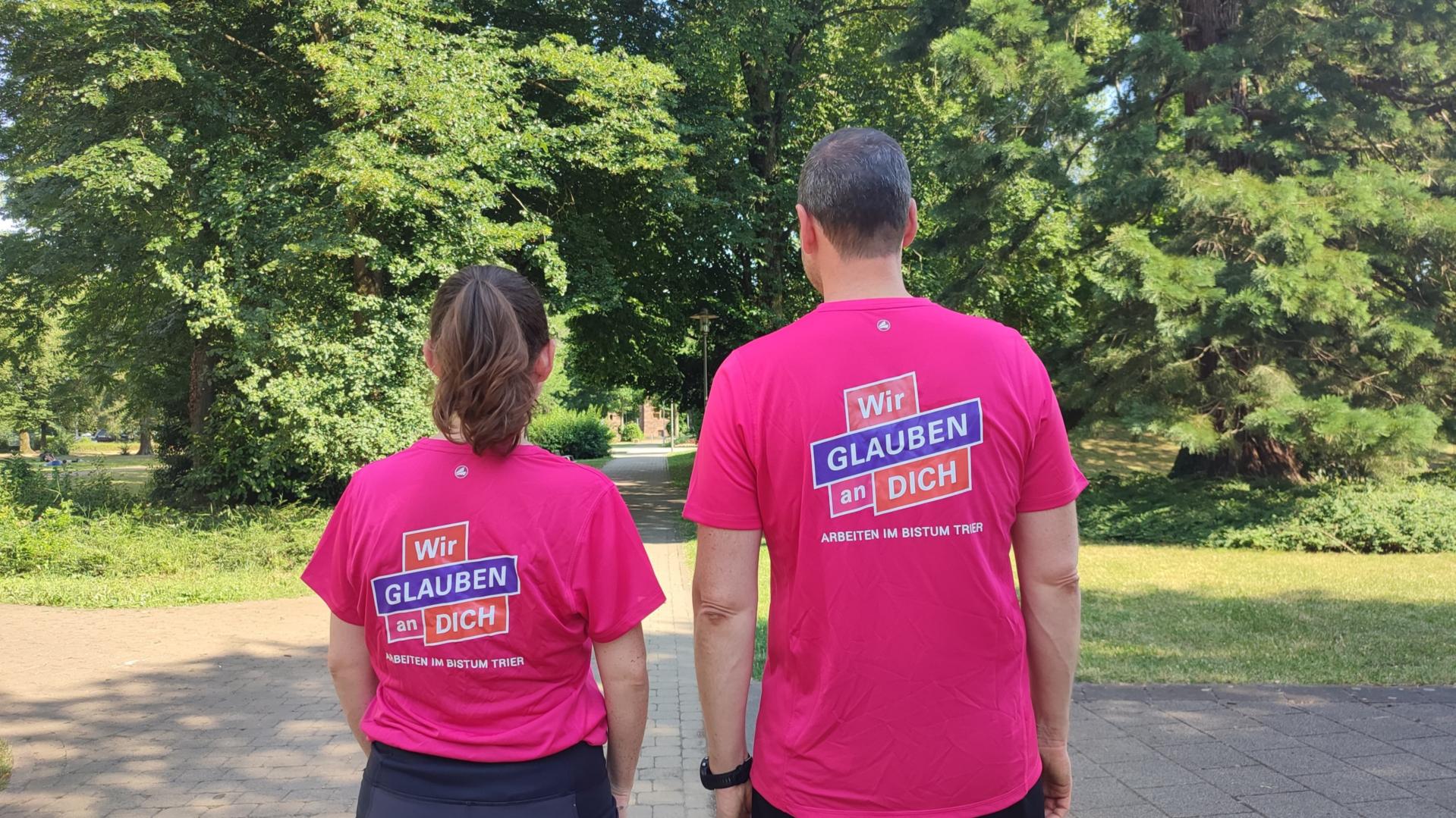 Zwei Personen stehen mit dem Rücken zur Kamera. Auf den Shirts steht wir glauben an dich.