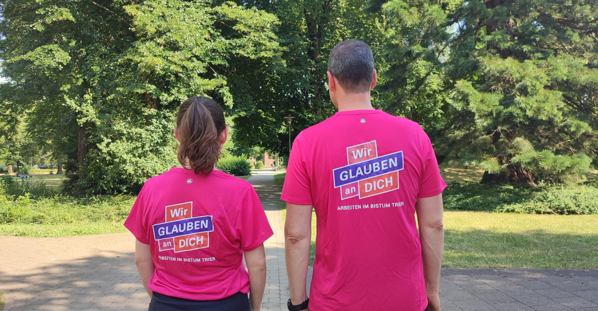 Zwei Personen stehen mit dem Rücken zur Kamera. Auf den Shirts steht wir glauben an dich.