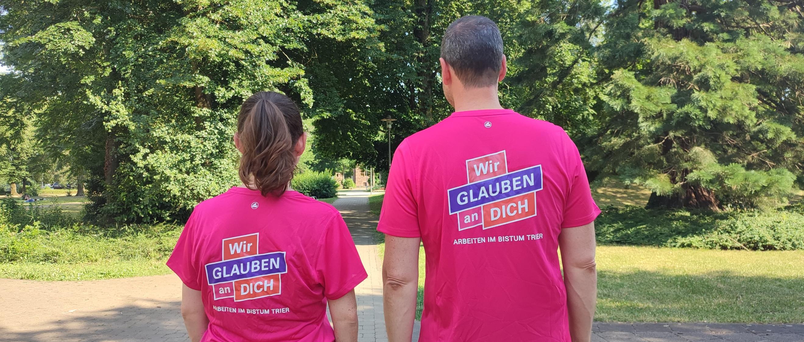 Zwei Personen stehen mit dem Rücken zur Kamera. Auf den Shirts steht wir glauben an dich.